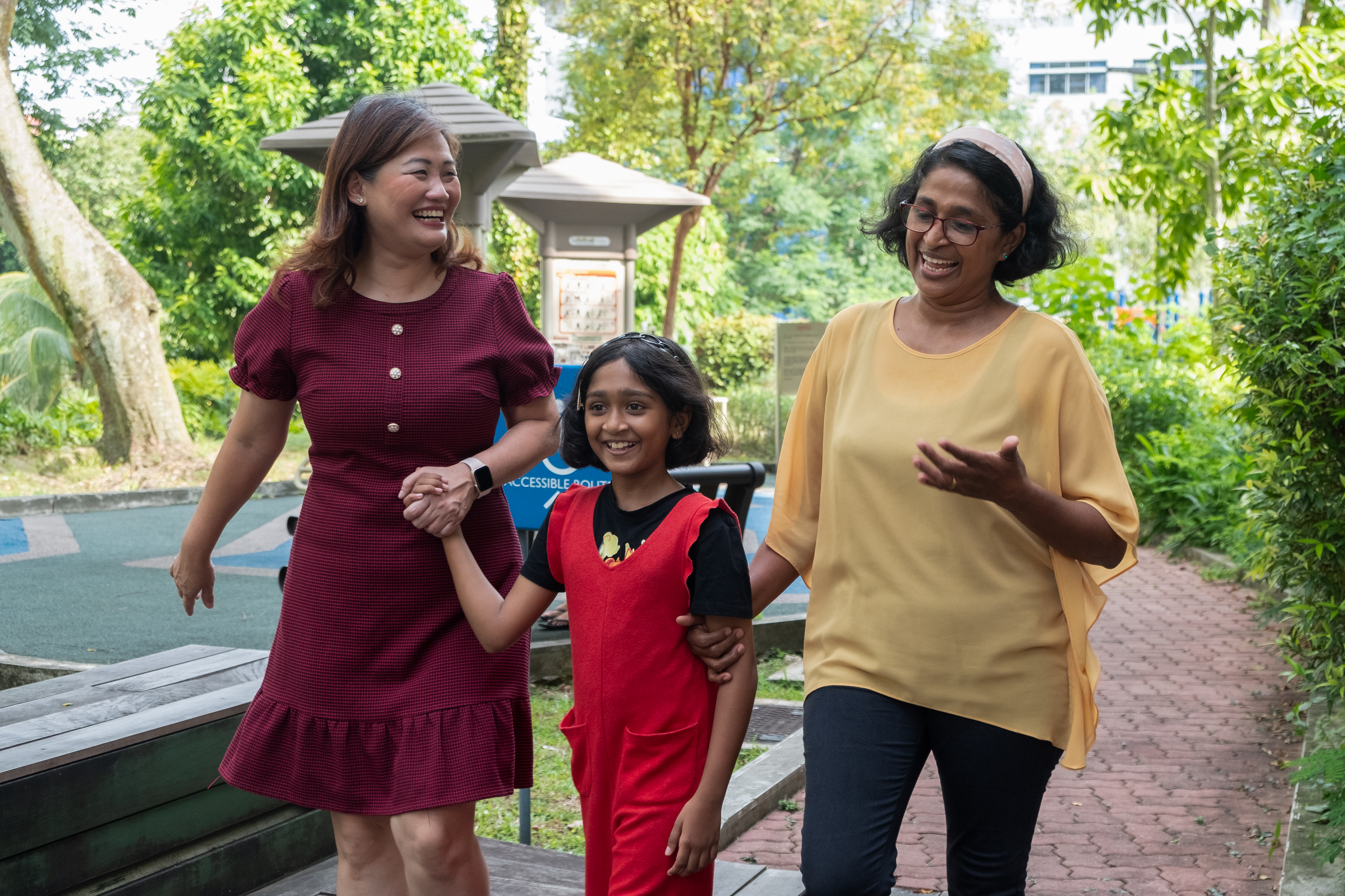 Cynthia, Upeka, and her daughter stroll down a path, holding hands and smiling while engaged in conversation amidst lush greenery.